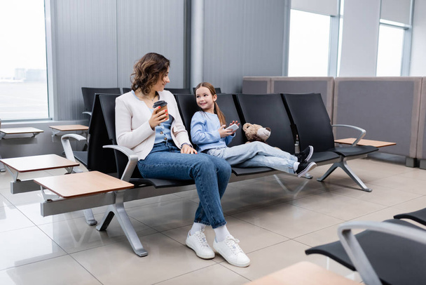 cheerful kid holding smartphone and looking at mother with paper cup in lounge hall of airport - Foto, immagini