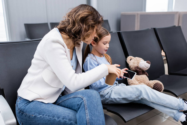 дитина вказує на смартфон поруч з матір'ю з паперовою чашкою в лаунж-залі аеропорту
  - Фото, зображення