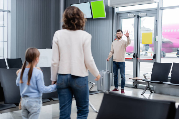 ευτυχισμένος άνθρωπος με αποσκευές κουνώντας το χέρι, ενώ κοιτάζοντας τη γυναίκα και την κόρη στο αεροδρόμιο  - Φωτογραφία, εικόνα