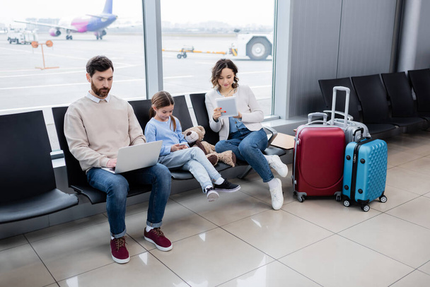 οικογένεια χρησιμοποιώντας συσκευές, ενώ κάθεται κοντά σε αποσκευές στο σαλόνι του αεροδρομίου  - Φωτογραφία, εικόνα