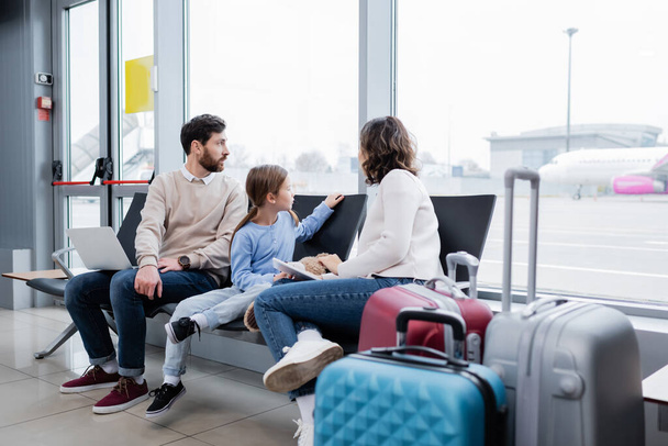 parents holding devices while sitting near daughter and looking at plane through window in airport lounge - 写真・画像