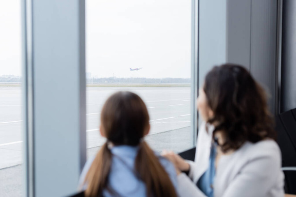 Mutter und Tochter blicken verschwommen durch Flughafenfenster auf startendes Flugzeug - Foto, Bild