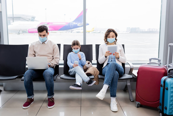 οικογένεια με ιατρικές μάσκες χρησιμοποιώντας gadgets στο αεροδρόμιο - Φωτογραφία, εικόνα