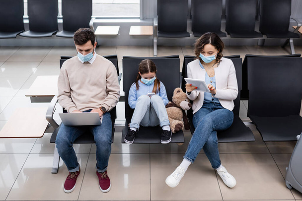 niño y padres en máscaras médicas usando aparatos en el aeropuerto - Foto, imagen