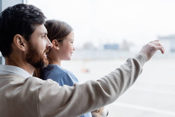 Glücklicher Vater zeigt auf Fenster neben lächelnder Tochter am Flughafen  - Foto, Bild
