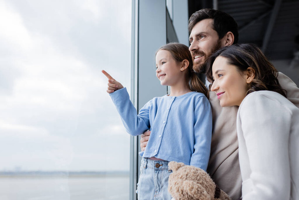 happy kid pointing with finger at window near cheerful parents in airport - Photo, Image