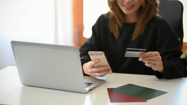Una joven mujer sosteniendo un teléfono inteligente y una tarjeta de crédito en su escritorio en la sala de estar. Pagar concepto de facturas. cosechados - Foto, Imagen