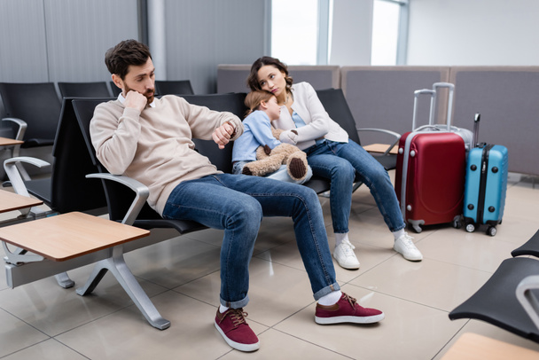 γενειοφόρος άνδρας κοιτάζοντας ρολόι κοντά στην κόρη και βαριέται σύζυγος στο σαλόνι του αεροδρομίου - Φωτογραφία, εικόνα