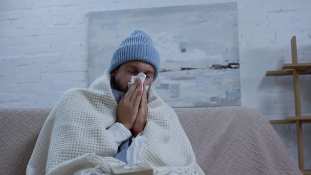 άρρωστος άνθρωπος σε ζεστό σκουφί και κουβέρτα φτάρνισμα σε χαρτοπετσέτα, ενώ κάθεται στον καναπέ - Φωτογραφία, εικόνα