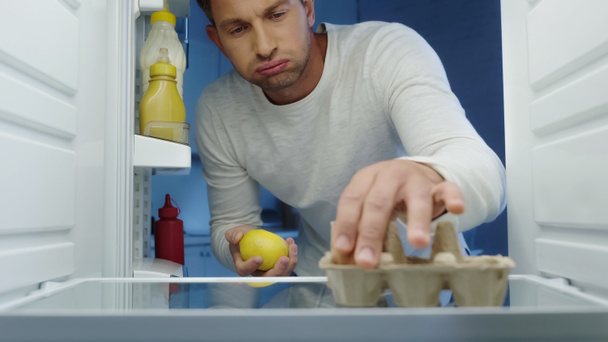 unhappy man puffing cheeks while taking egg and lemon from fridge - Fotó, kép