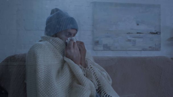 sick man sitting on couch in warm beanie and blanket and sneezing in paper napkin - Fotó, kép