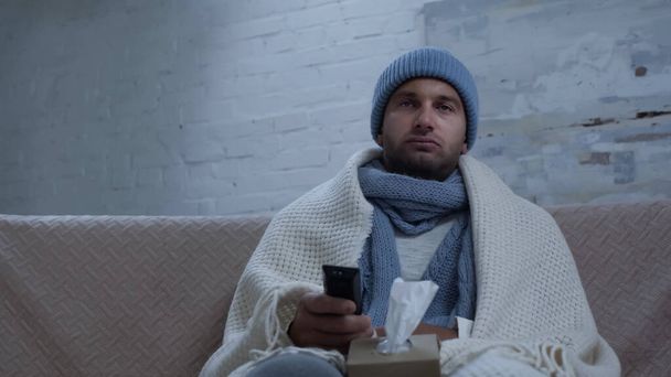 άρρωστος άνθρωπος με τηλεχειριστήριο τηλεόρασης και χαρτοπετσέτες κάθεται στον καναπέ σε ζεστό σκούφο και κουβέρτα - Φωτογραφία, εικόνα