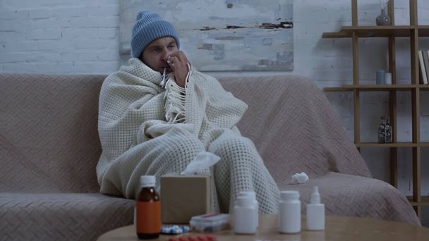 Hastalıklı bir adam, sıcak bir şapka ve battaniyeyle kanepeye oturmuş ilaçla burnunu kağıt peçeteyle siliyor. - Fotoğraf, Görsel