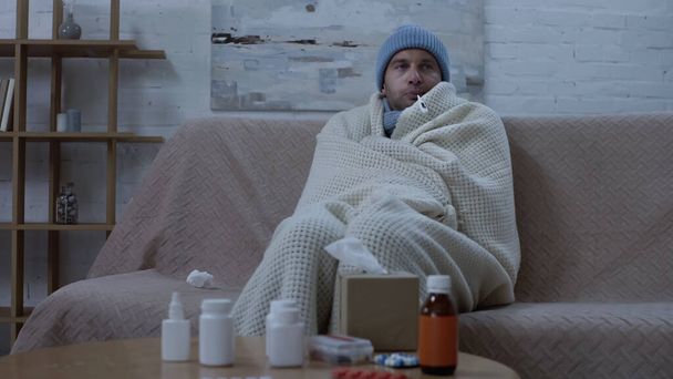 хворий чоловік вимірює температуру, сидячи на дивані в теплих бджіл і ковдрах біля столу з ліками
 - Фото, зображення