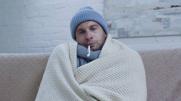 άρρωστος άνθρωπος μέτρηση της θερμοκρασίας, ενώ κάθεται στον καναπέ σε ζεστό σκούφο και κουβέρτα - Φωτογραφία, εικόνα