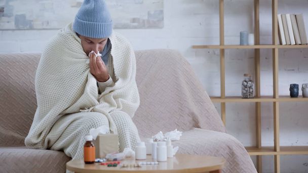 ill man wiping nose with paper napkin while sitting in warm blanket and hat near medicaments on table - Foto, imagen