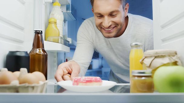 happy man taking delicious doughnut from fridge near beverages, eggs, bottle and jars with food - 写真・画像