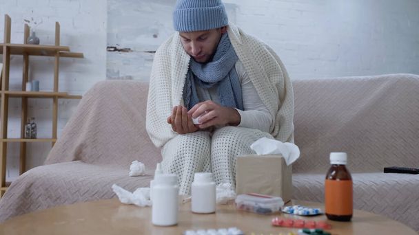 sick man in warm blanket and hat holding pills container near table with medicaments - Zdjęcie, obraz