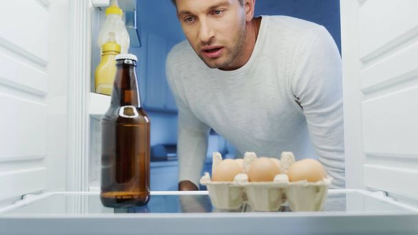 exhausted man looking at bottle of beer in fridge near eggs and sauces - Foto, immagini