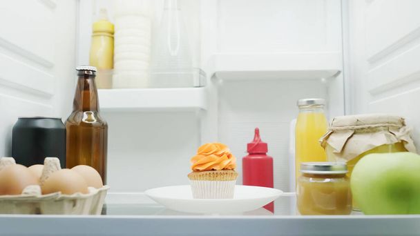 sabroso cupcake cerca de los huevos, botella con bebidas, frascos con miel y puré de frutas en nevera - Foto, Imagen