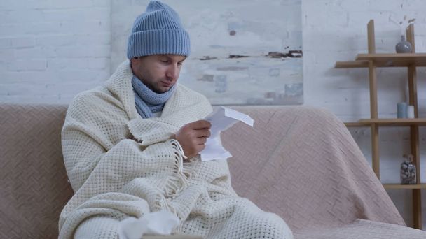 Sıcak bereli ve battaniyeli hasta adam evdeki kanepede ilaç dersleri okuyor. - Fotoğraf, Görsel