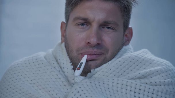 upset diseased man in blanket looking at camera while measuring temperature with thermometer in mouth - Photo, Image