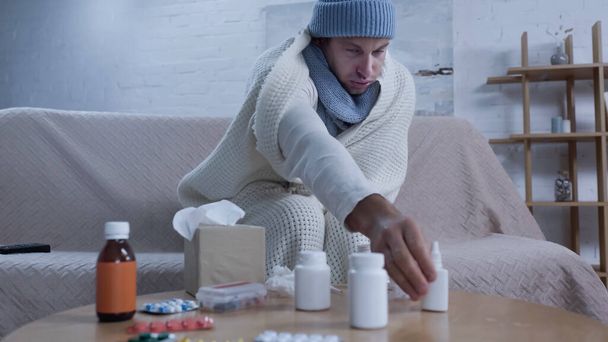 Sıcak battaniyeli ve şapkalı hasta adam burun spreyine uzanırken ilaçların yanında kanepede oturuyor. - Fotoğraf, Görsel