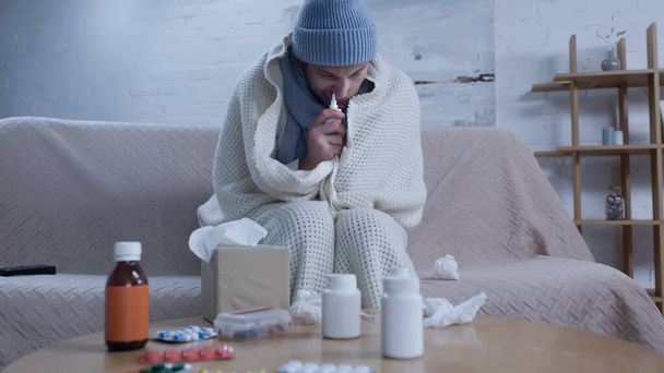 diseased man in warm blanket and beanie using nasal spray while suffering from runny nose near table with medication - Photo, Image