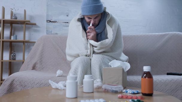 ill man using nasal spray while sitting in warm beanie and blanket near medicaments and crumpled paper napkins - Fotografie, Obrázek