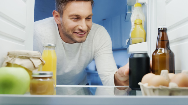 happy man taking soda from fridge near bottle of beer, orange juice, eggs, and jars with food - Photo, Image