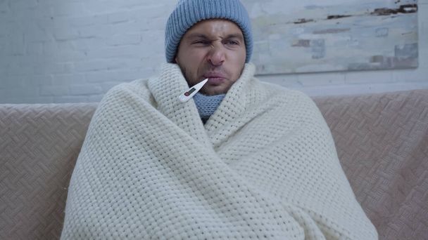 Sıcak şapkalı ve battaniyeli hoşnutsuz hasta bir adam. Ağzında termometreyle ateşi ölçüyor. - Fotoğraf, Görsel