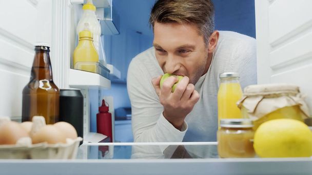 Adam buzdolabının yanında yumurtalarla, içeceklerle, konteynırlarla ve yiyeceklerle olgun elma yiyor. - Fotoğraf, Görsel