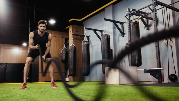 full length of strong sportsman training with battle ropes in gym - Foto, Imagem