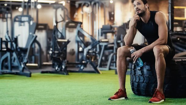 tired sportsman in earphone resting on car tire and holding sports bottle in gym  - Photo, image