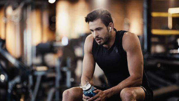 tired sportsman in earphone listening music and holding sports bottle in gym  - Photo, Image