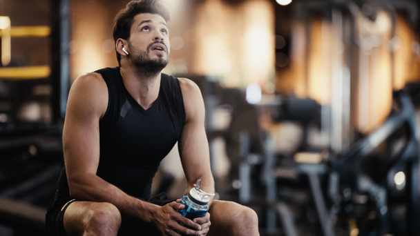 tired sportsman in earphone listening music, resting and holding sports bottle in gym  - Photo, image
