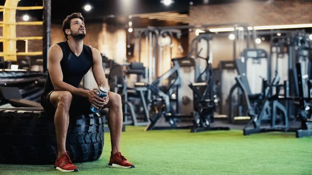 exhausted man in earphone listening music, sitting on car tire and holding sports bottle in gym - Photo, Image