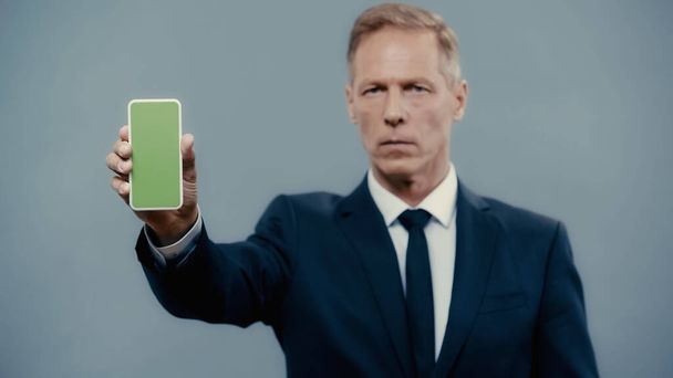 Smartphone with green screen in hand of blurred businessman isolated on grey  - Фото, изображение