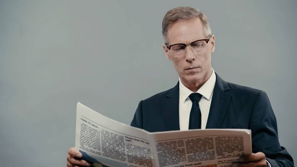 Mature businessman in suit and eyeglasses reading newspaper isolated on grey  - Fotó, kép