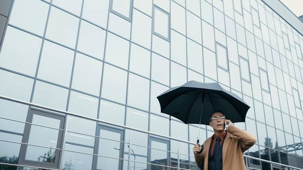 Empresario sosteniendo paraguas mientras habla en el teléfono inteligente y caminando cerca del edificio al aire libre  - Foto, imagen