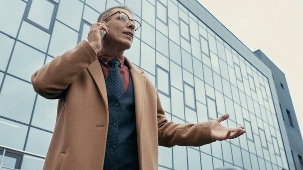 Low angle view of businessman in coat talking on cellphone near building on urban street  - 写真・画像
