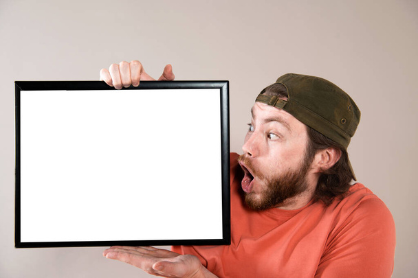 Young bearded man making funny face, holding a blank sign board. Studio shot on gray background. - Photo, Image
