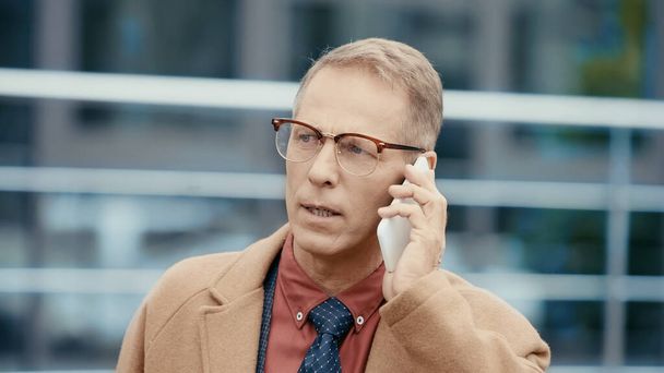 Middle aged businessman in eyeglasses and coat talking on cellphone outdoors  - Photo, Image