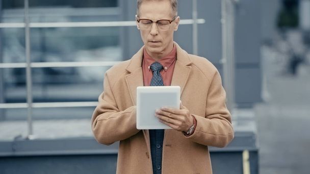 Mature businessman in coat using digital tablet on urban street  - 写真・画像