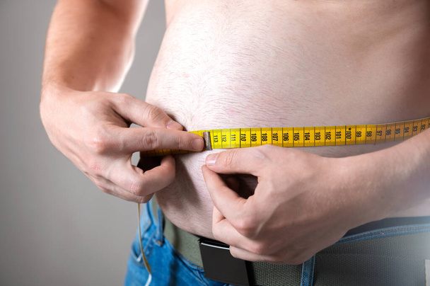 Un hombre mide su vientre gordo con una cinta métrica sobre un fondo gris - Foto, Imagen