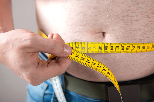 A man measures his fat belly with a measuring tape on a gray background - Photo, Image