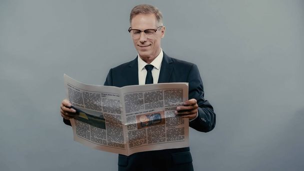Smiling businessman in eyeglasses reading newspaper isolated on grey  - Fotó, kép