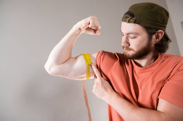 Vousatý mladík měřící biceps, svaly na paži žlutou páskou. - Fotografie, Obrázek