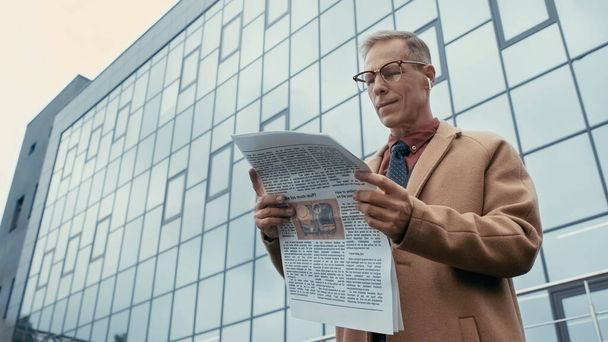 Low angle view of businessman in earphone and coat reading newspaper on urban street  - Photo, image