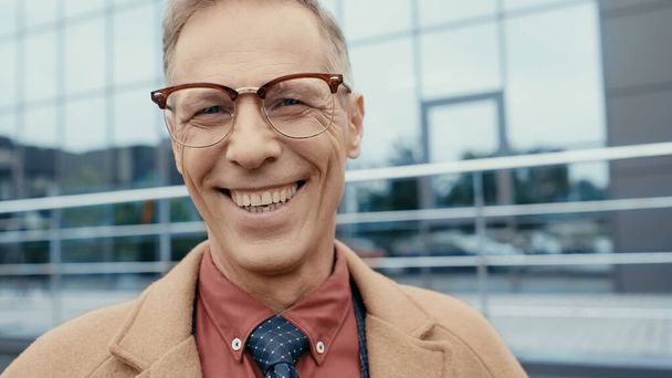 Portrait of happy businessman in coat and eyeglasses looking at camera outdoors  - Foto, afbeelding
